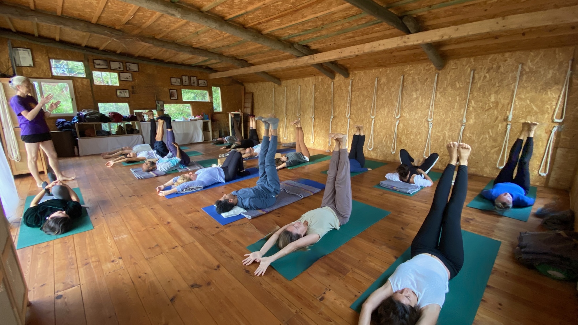 Students practicing legs-up-the-wall pose in Georgia's rustic wooden studio in Colares, Portugal, with exposed beams and traditional Iyengar yoga rope props on the walls