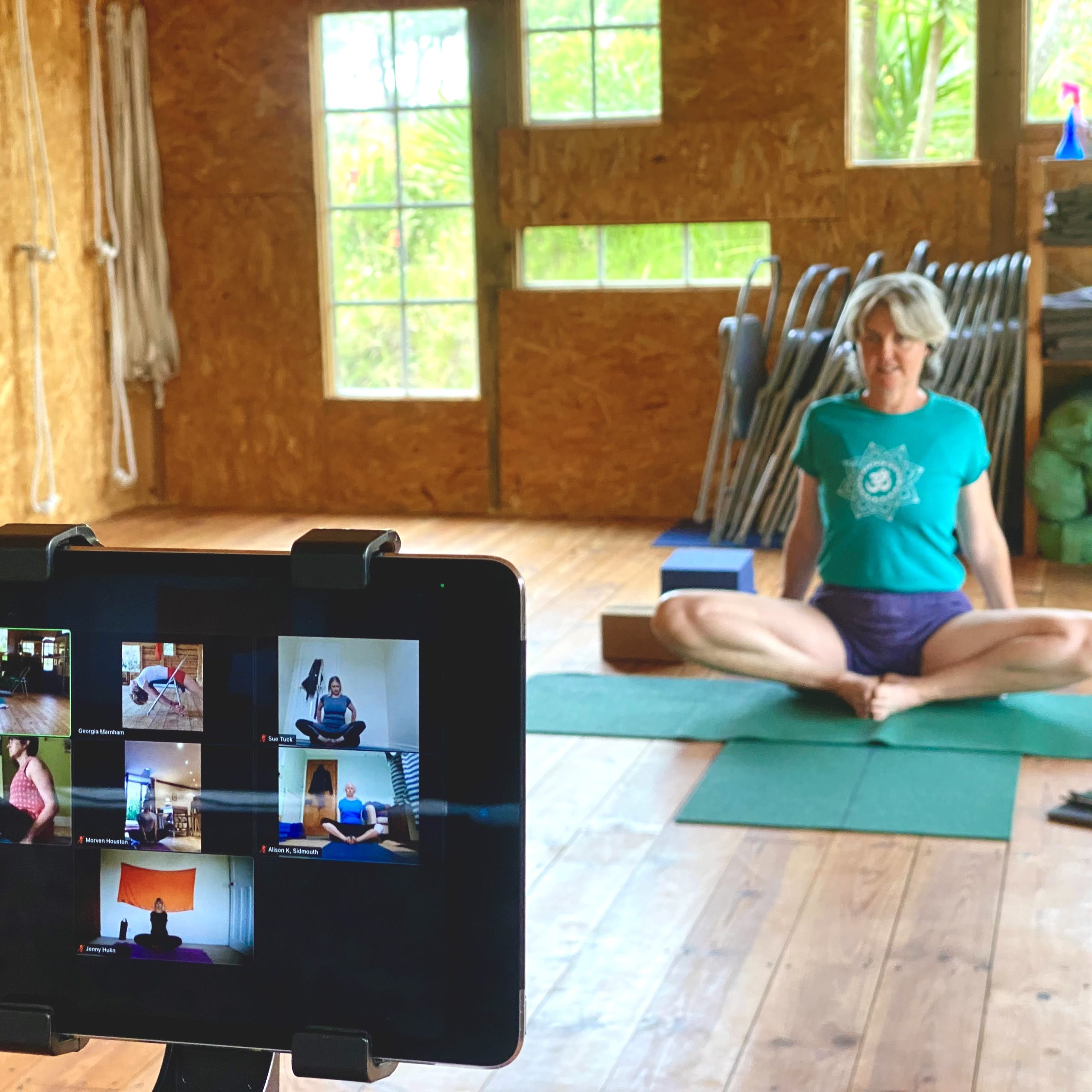 Georgia teaching an online Iyengar Yoga class from her studio in Portugal, sitting in meditation pose on a green mat while students join via Zoom on a tablet screen, showing the authentic online teaching setup