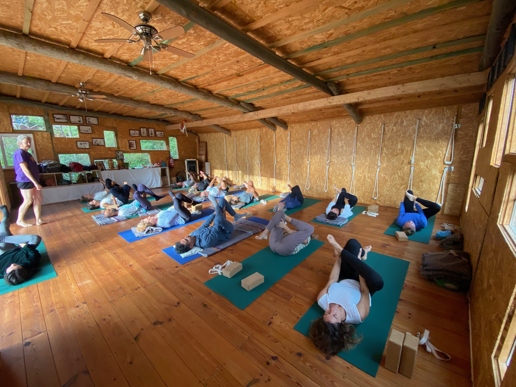Georgia teaching an Iyengar Yoga class in her rustic wooden studio in Colares, Portugal, with students practising restorative poses on green and blue mats, showing the authentic studio atmosphere with exposed beams and traditional rope props
