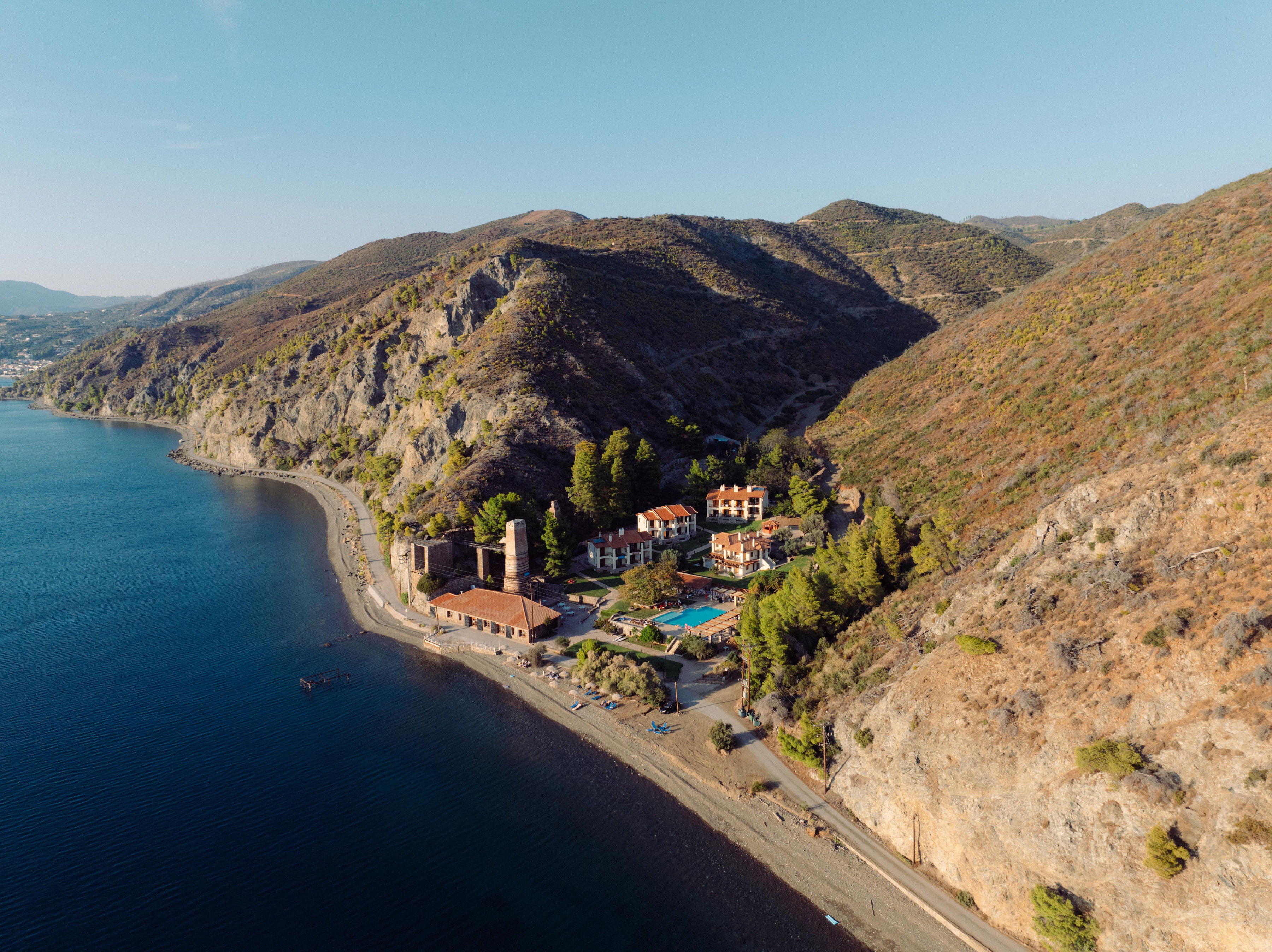 Aerial view of Kaminos Resort nestled against dramatic hills with the Aegean Sea coastline, showing terracotta roofs, swimming pool, and pine forests