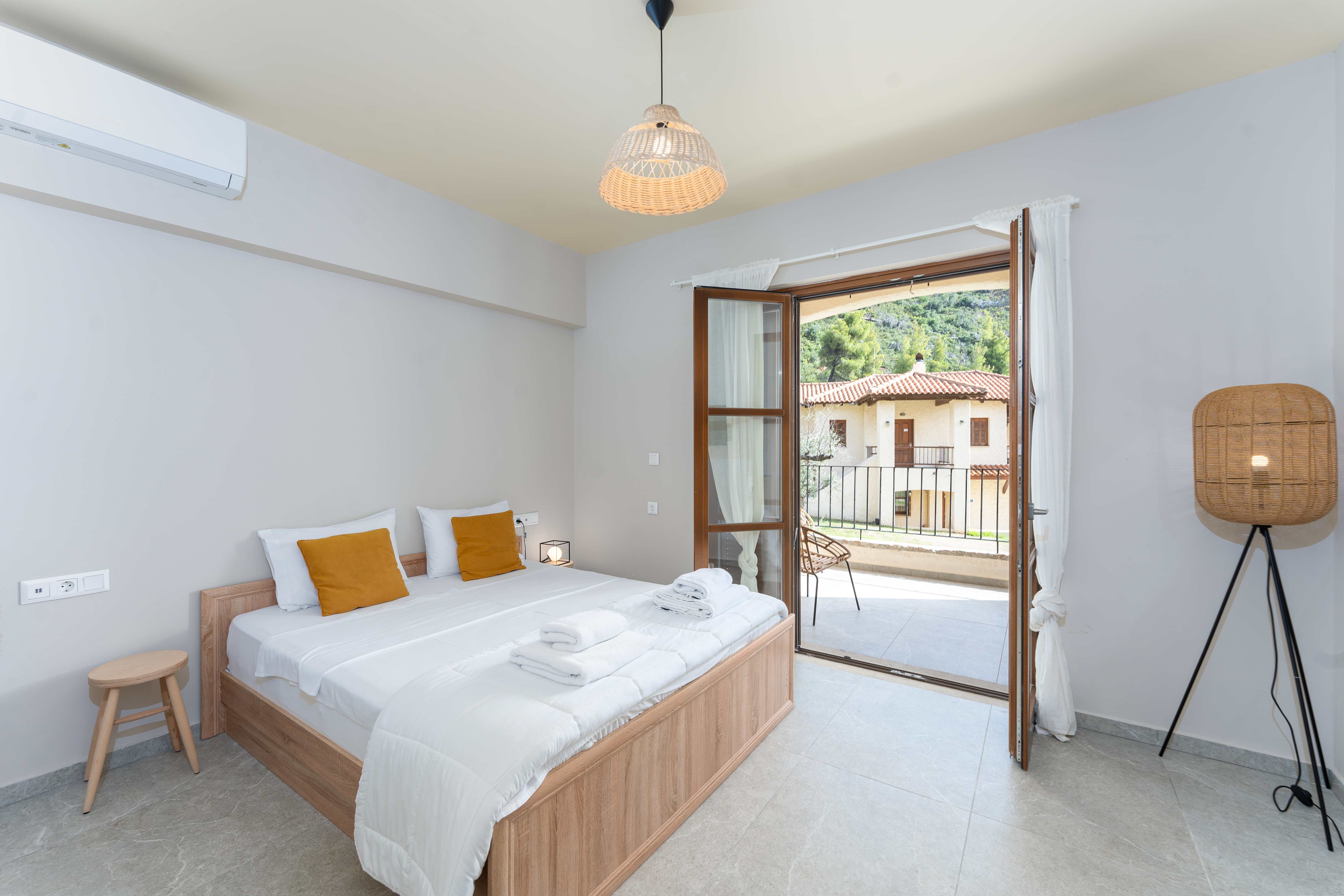 Modern bedroom at Kaminos Resort with wooden furniture, wicker lighting, and balcony doors opening to views of Mediterranean buildings and hills