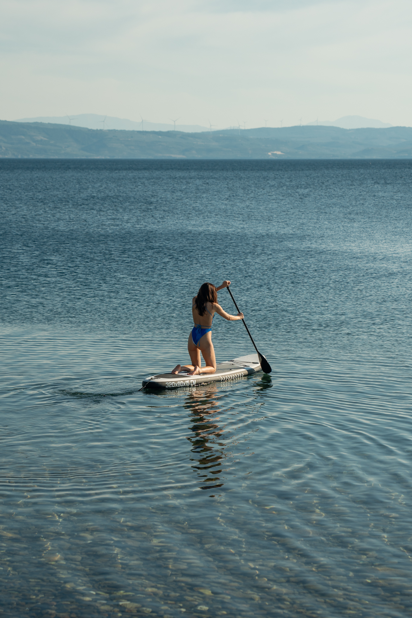 Woman paddleboarding on calm Aegean Sea waters during golden hour with distant hills on the horizon