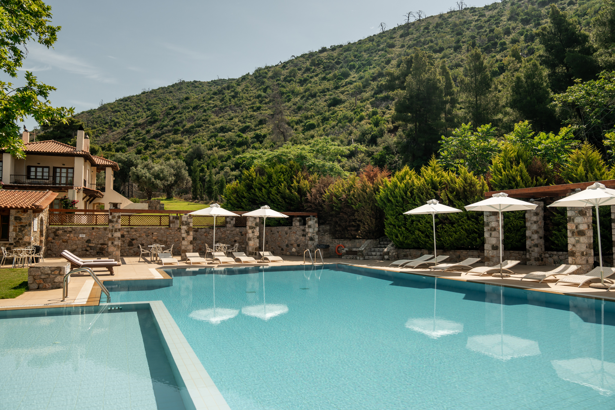 Large swimming pool at Kaminos Resort with turquoise water, white umbrellas, sun loungers, stone pergolas, and lush green hills in the background