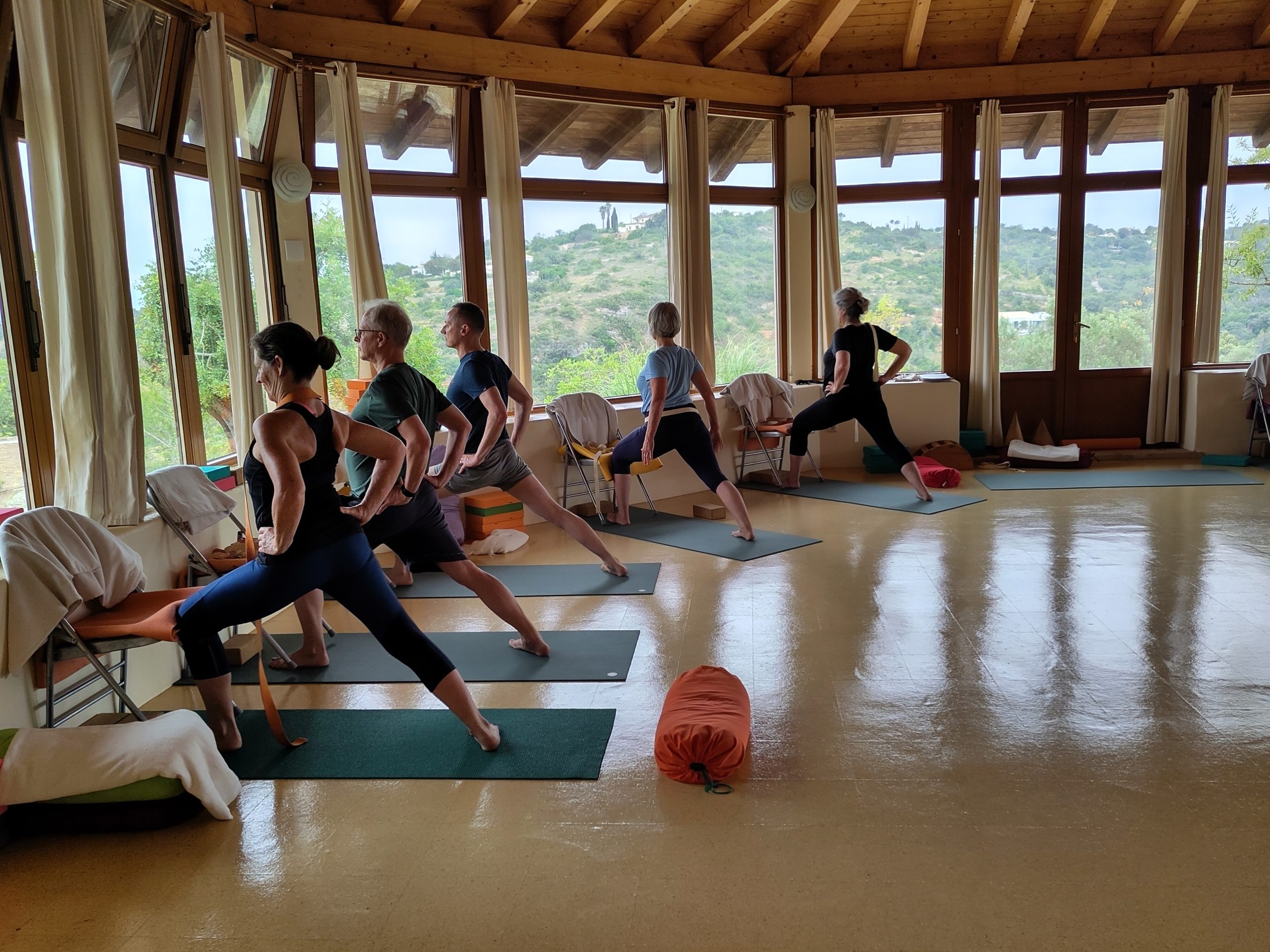 Students practising Iyengar Yoga in a beautiful retreat studio with floor-to-ceiling windows overlooking rolling countryside, using traditional props like chairs and blocks in a spacious, light-filled room with exposed wooden beams